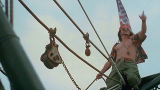 Gary Sinise with an open shirt, atop a the mast of a boat with an American flag behind him in Forrest Gump