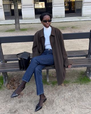 Influencer Sylvie Mus sits on a park bench in Paris wearing oval sunglasses, a white turtleneck top, brown coat, dark-wash jeans, black sheer tights, and black kitten pumps.