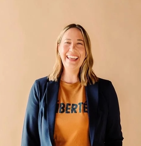 A smiling blonde woman with an orange shirt and blue blazer.