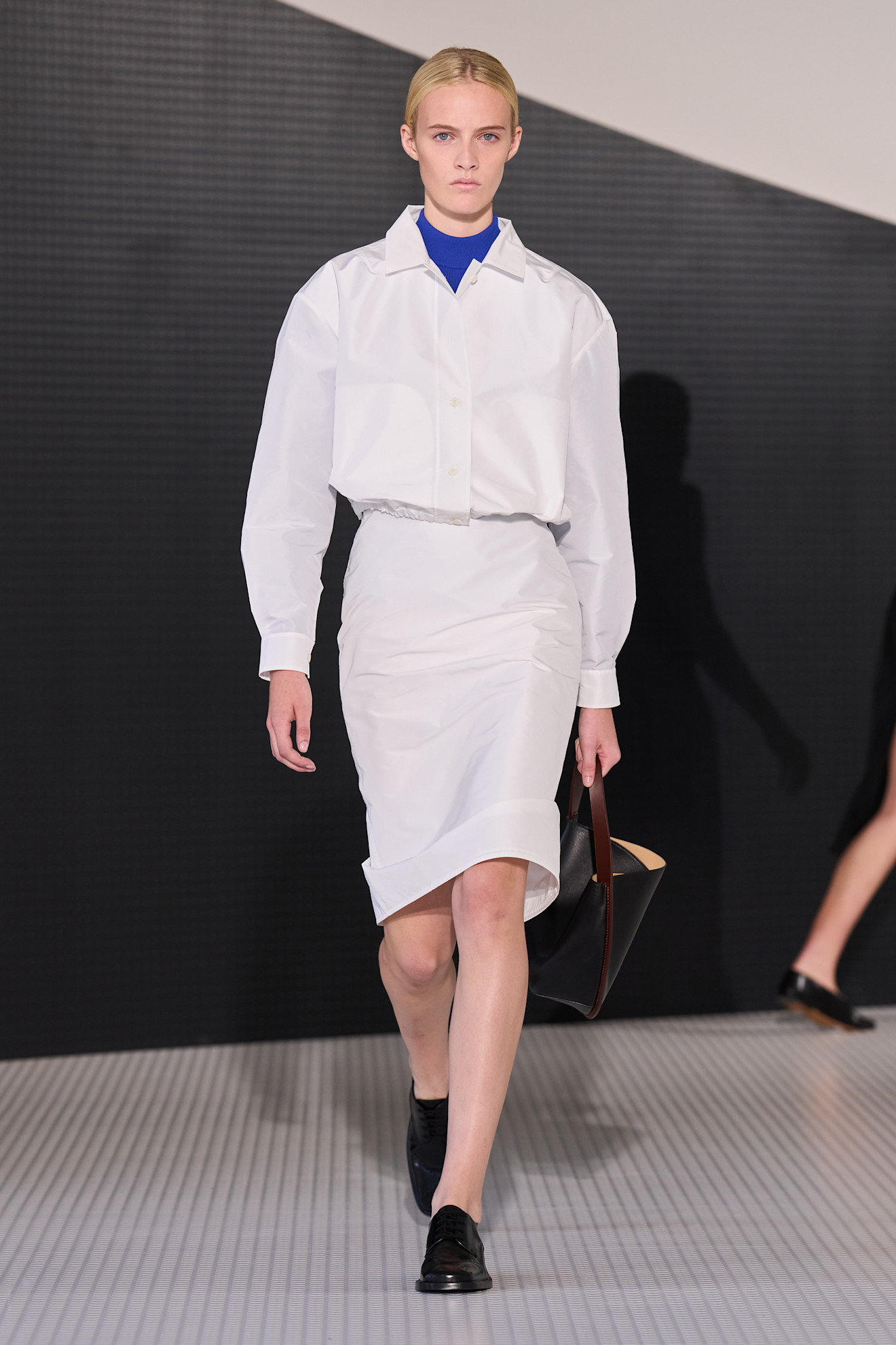 Layering staples for spring 2026 is shown in a photo from Jil Sander's runway show, with a model walking while wearing a cobalt blue turtleneck under a white button-down shirt styled with a white pencil skirt, black loafers, and a black handbag