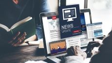 A man looking at news in various format on a laptop, with graphics popping out of the screen, while a woman opposite him reads a book