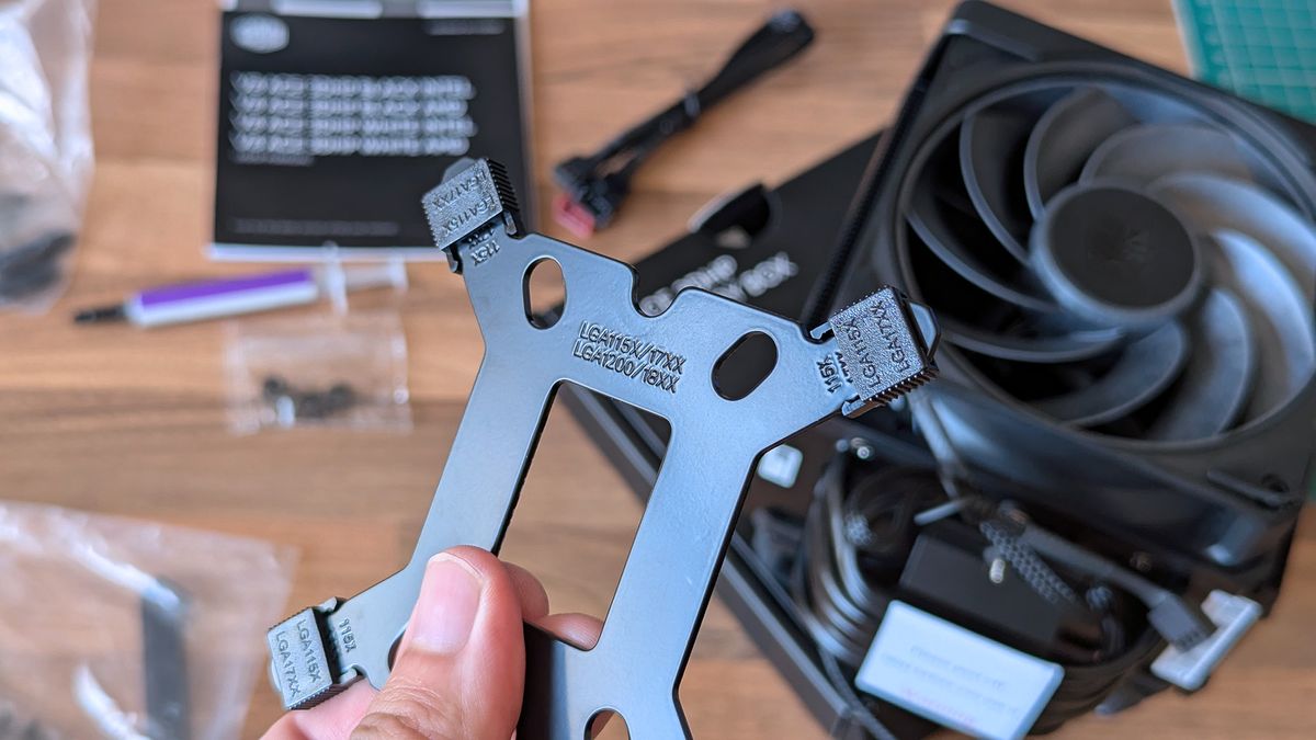 A hand holds a metal CPU cooler mounting bracket labeled for Intel LGA sockets. Screws, cables, a fan, and thermal paste are scattered across the desk behind it.