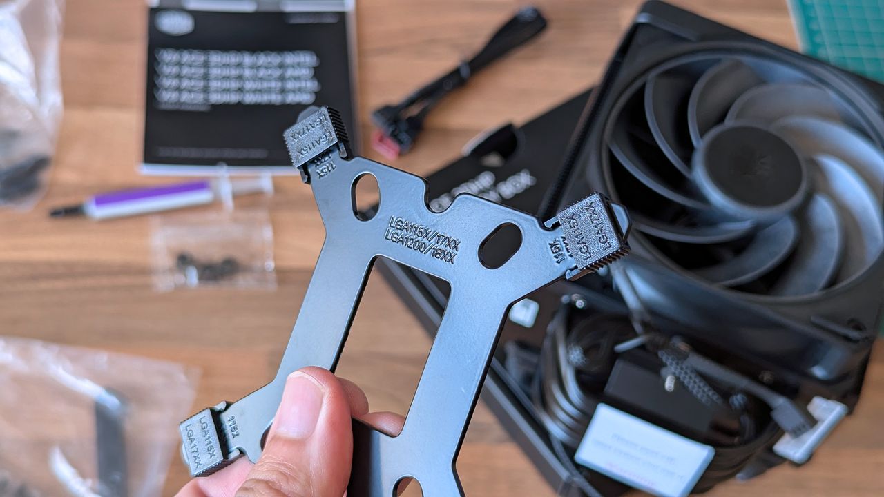A hand holds a metal CPU cooler mounting bracket labeled for Intel LGA sockets. Screws, cables, a fan, and thermal paste are scattered across the desk behind it.