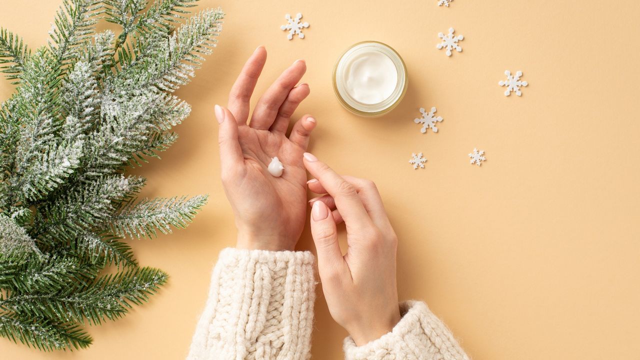 girl&#039;s hands in white sweater applying cream on her hands from jar fir branches in frost and snowflakes on isolated pastel beige background 