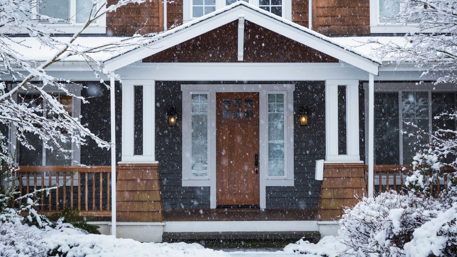 Exterior shot of a large home in the winter, with snow on peaked porch, front yard, path and falling. The front door is wooden as are the porch guard rails