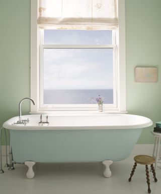 A bathroom with light green walls, a window overlooking the sea, and a white standing bath tub with a wooden stool next to it