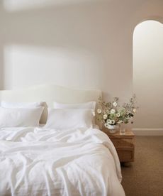 White 100% French Flax Linen Bedding Set on a bed against a cream wall; to the right, a wooden nightstand topped with a vase of flowers. 