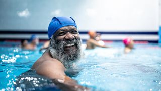 older man with greyin beard wears a swim cap and smiles as he's swimming at a public pool