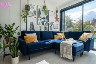 A blue velvet sectional sofa for a small living room painted in white, with large windows, gallery wall and pot plants.