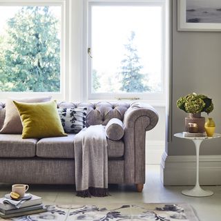 grey living room with floral rug, grey sofa with button back, tumeric cushion, grey fringed blanket, small round side table, vase, artwork
