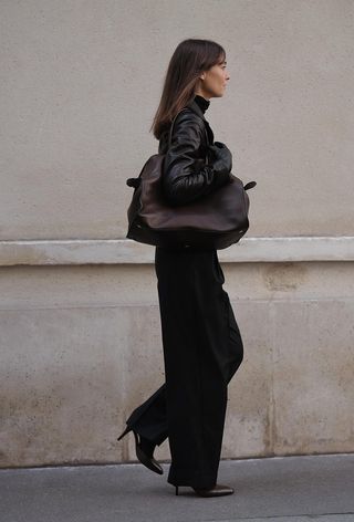 Black turtleneck outfit is shown in a photo of a French woman on the sidewalk wearing a black leather blazer over a black turtleneck with black leather gloves, a brown handbag, black trousers, and brown slingbacks