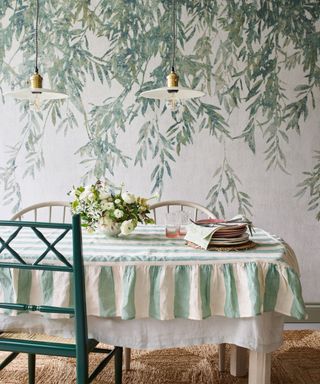 Dining room with textured, white and green botanical mural, white and green striped tablecloth on dining table, two traditional light wooden chairs, one painted green wooden dining chair,two low hanging glass pendants, table dressed with flowers, plates and glassware