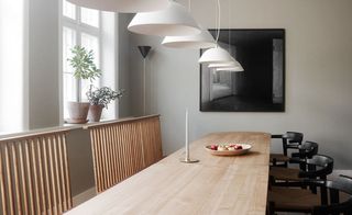 View of the dining area at The Apartment gallery featuring a long wooden dining table with a candle and a bowl of fruit on top, light wood and black chairs, grey walls, white pendant lights, framed wall art, a floor lamp and a window with plants in pots on the window sill