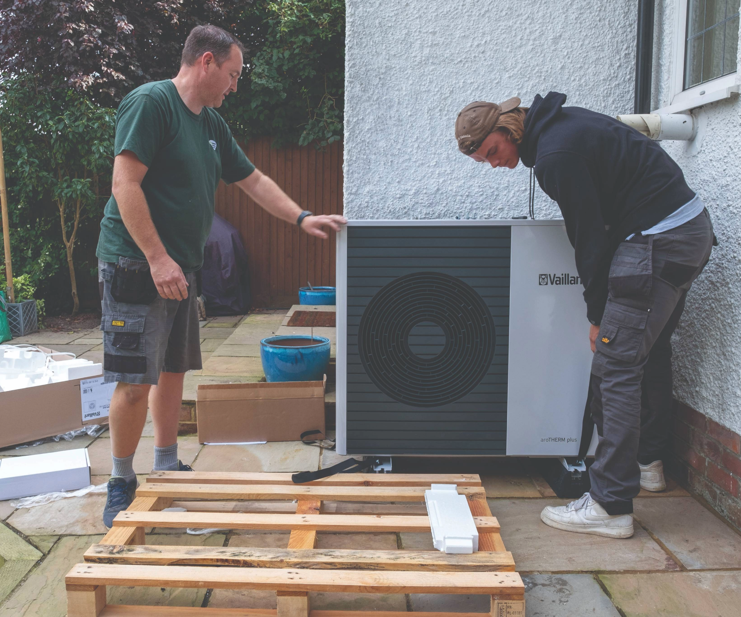 Two people holding a heat pump in the back garden of a house