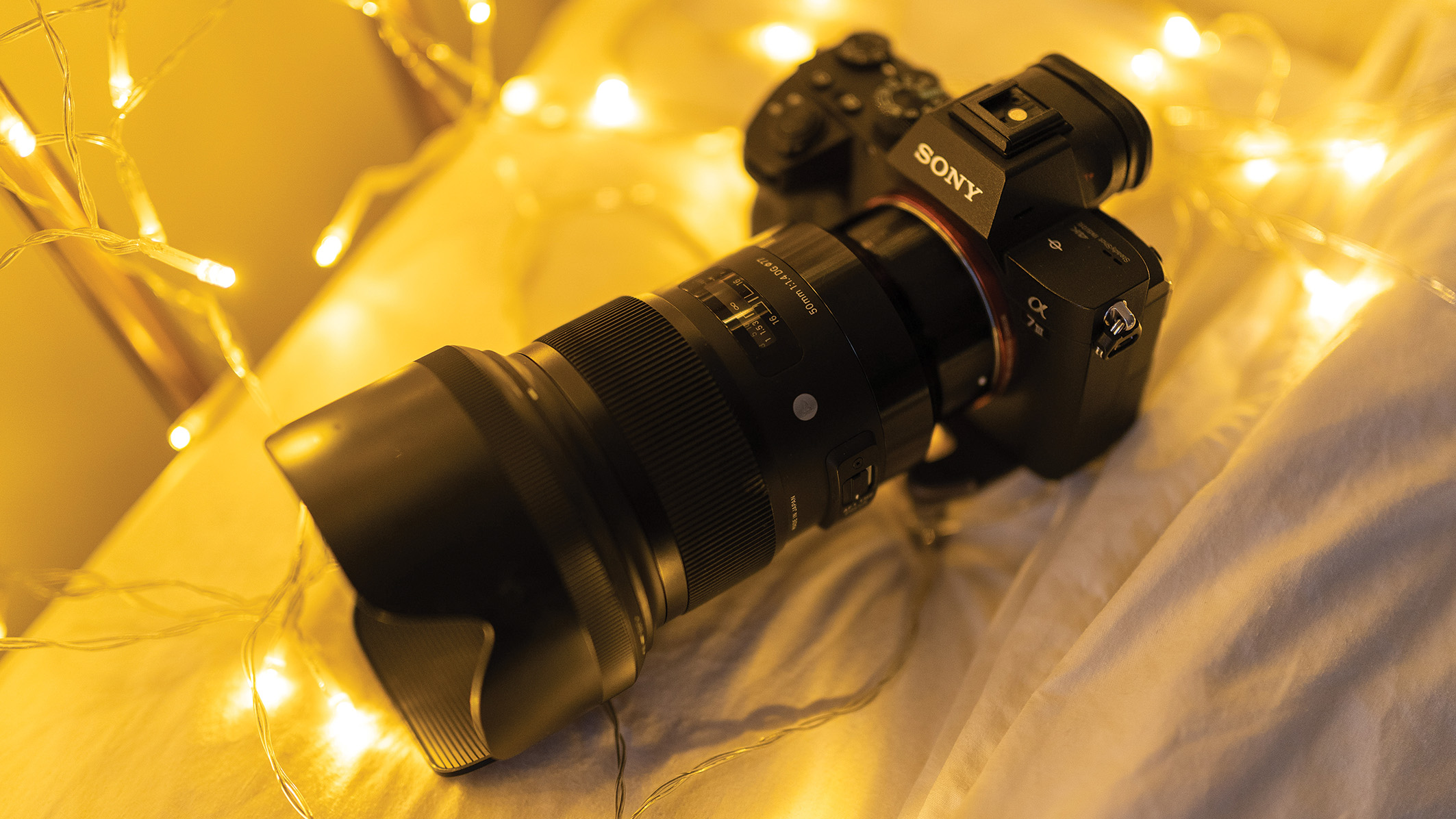 A Sony camera with a large lens rests on a bed surrounded by glowing fairy lights. The warm lighting creates a cozy, inviting atmosphere