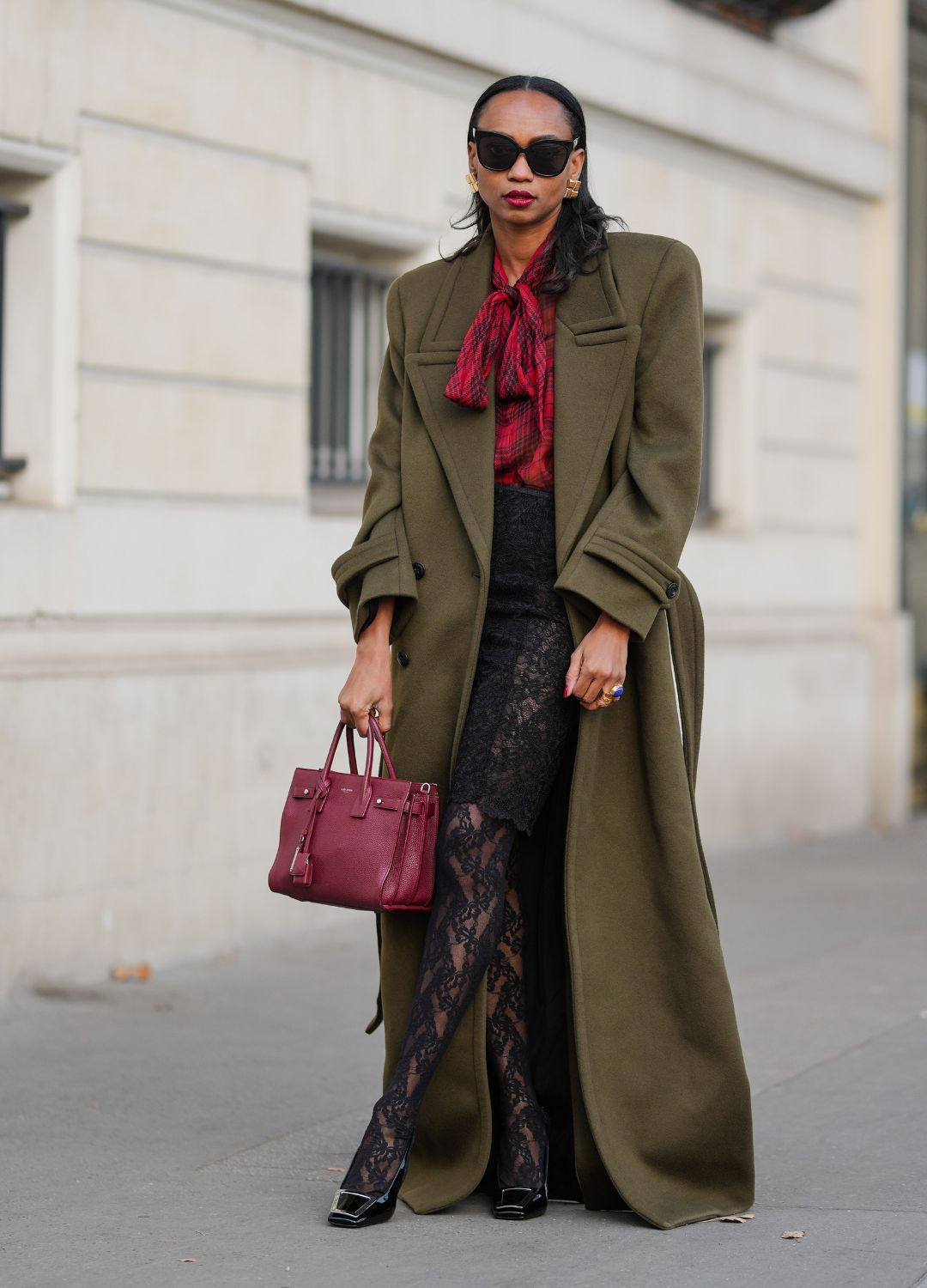 Emilie Joseph wears black sunglasses, gold earrings, a khaki long YSL trench coat, a red YSL shirt, a black lace skirt, gold rings, a burgundy leather Birkin Hermes bag, black lace tights, black leather high heels shoes, during a street style fashion photo session, on November 16, 2024 in Paris, France.
