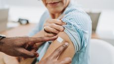 A photo shows a close-up on a doctor's hands as he applies a bandaid to an older woman's arm, as if she's just gotten a vaccine