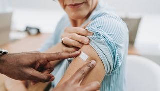 A photo shows a close-up on a doctor's hands as he applies a bandaid to an older woman's arm, as if she's just gotten a vaccine