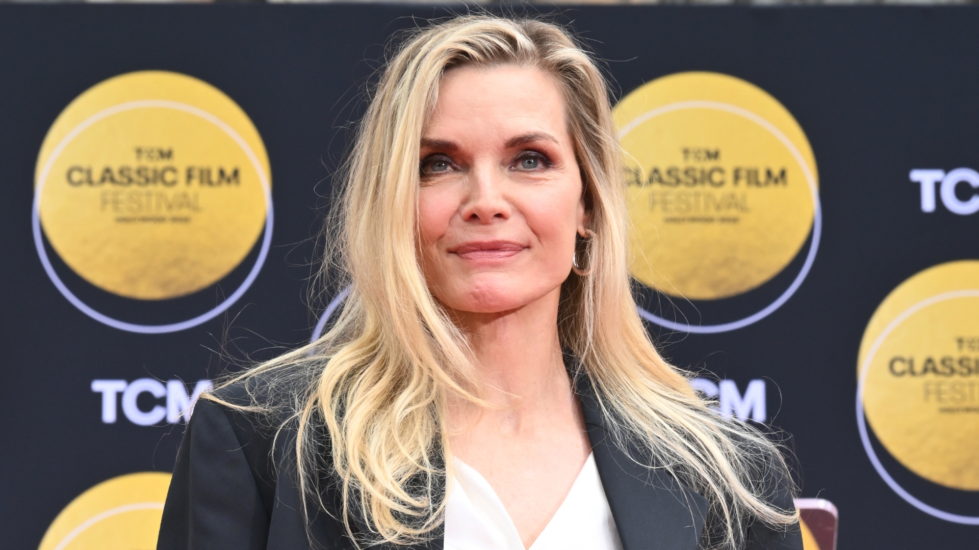 Michelle Pfeiffer is pictured with side-swept hair and wearing a black blazer and white silk shirt at the TCM ceremony honoring Michelle Pfeiffer with Hand and Footprint Ceremony as part of the TCM Classic Film Festival held at the TCL Chinese Theater on April 25, 2025 in Los Angeles, California.