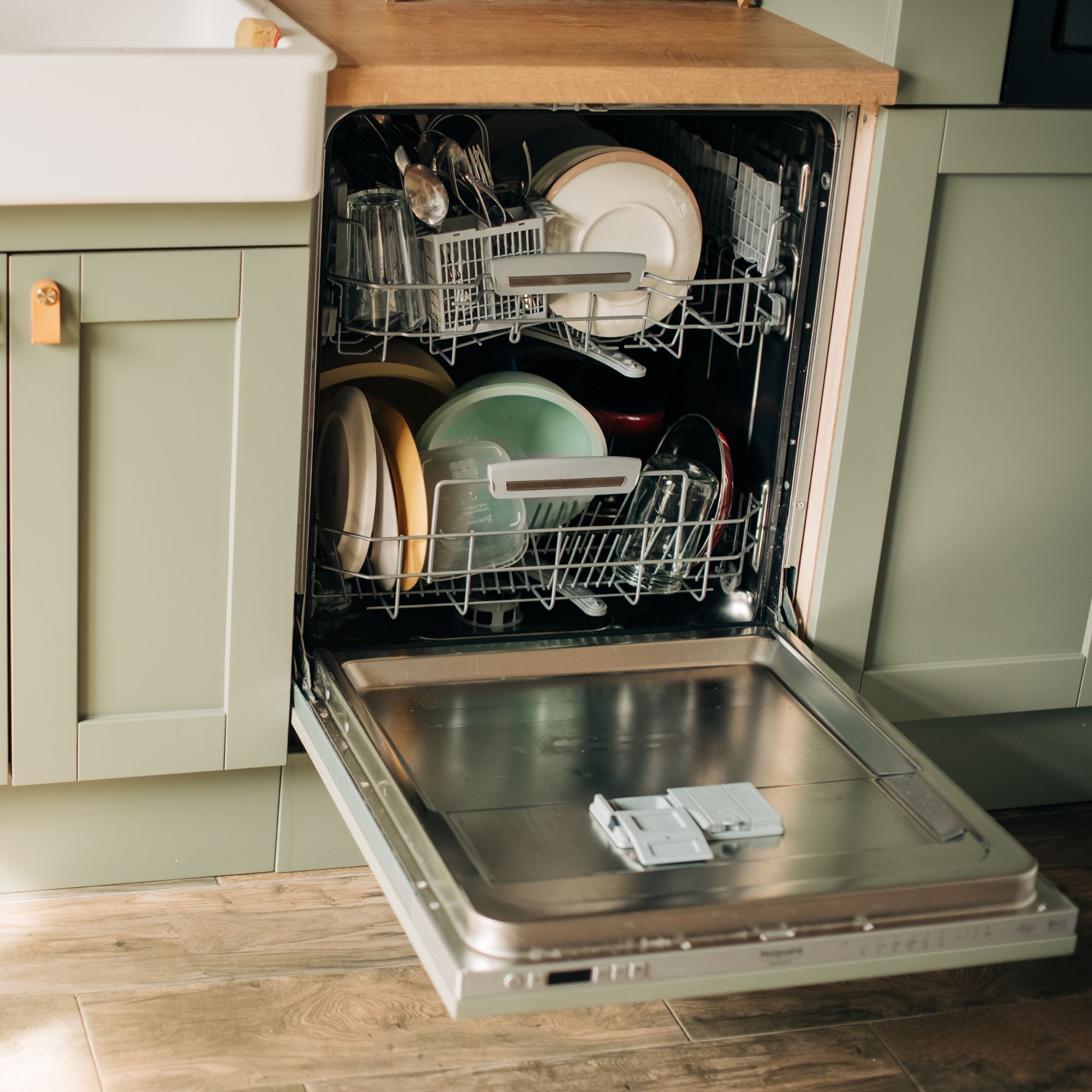 Open door of a loaded dishwasher in a kitchen with sage green cupboard doors, wood worktops and butler sink