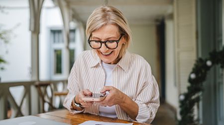 An older woman smiles as she looks at her phone while sitting at a table on her porch.