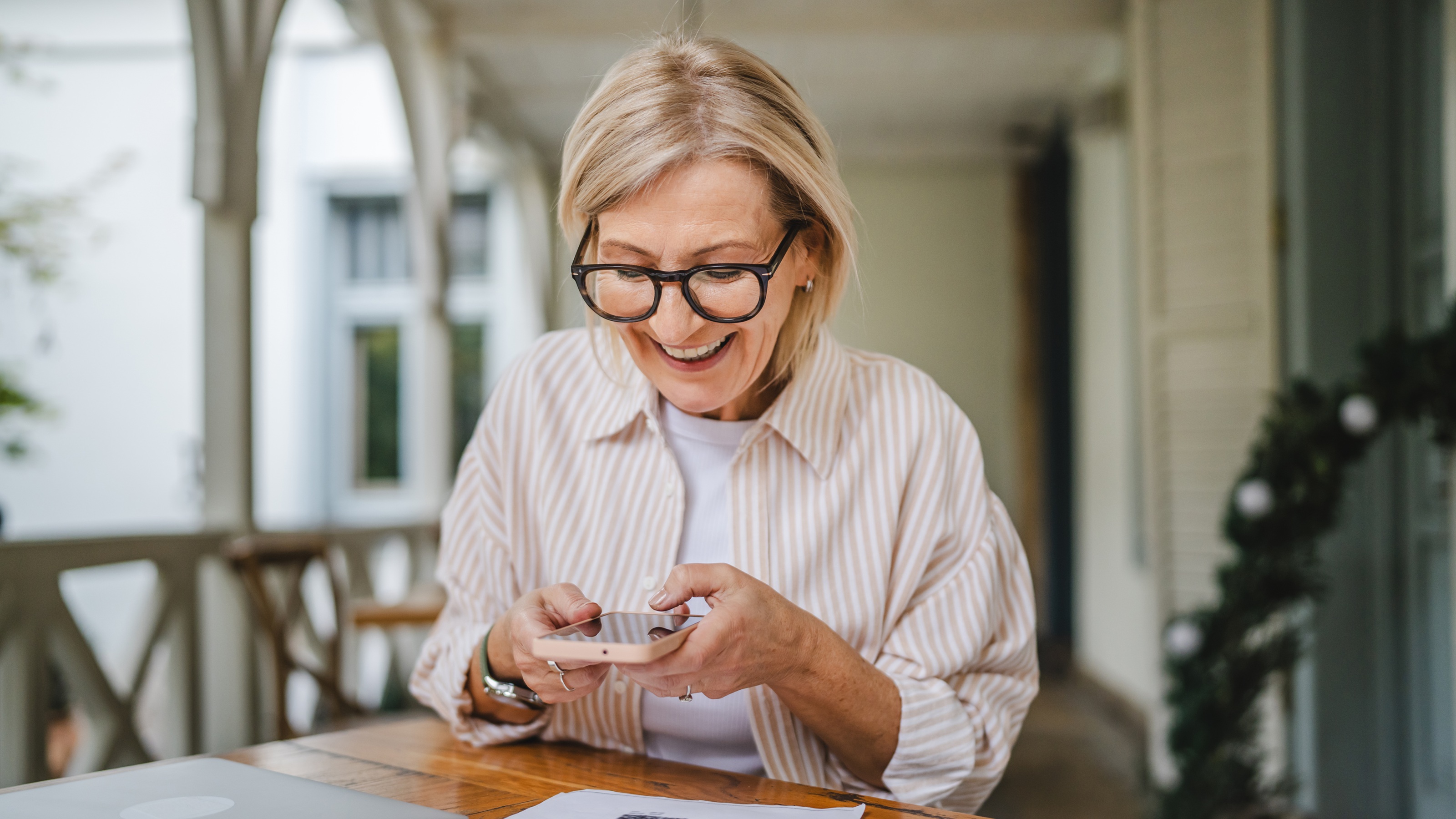 An older woman smiles as she looks at her phone while sitting at a table on her porch.