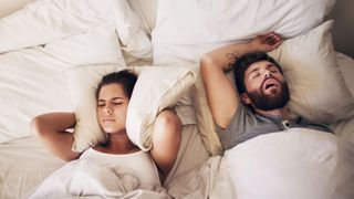 A man snores while asleep in bed as a woman next to him covers her ears with a pillow to block out the noise.