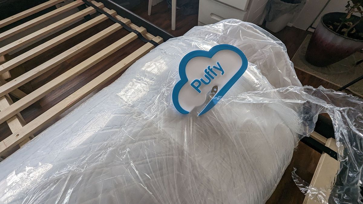 Puffy Lux Hybrid mattress vacuum packed and rolled in plastic, next to a bed frame