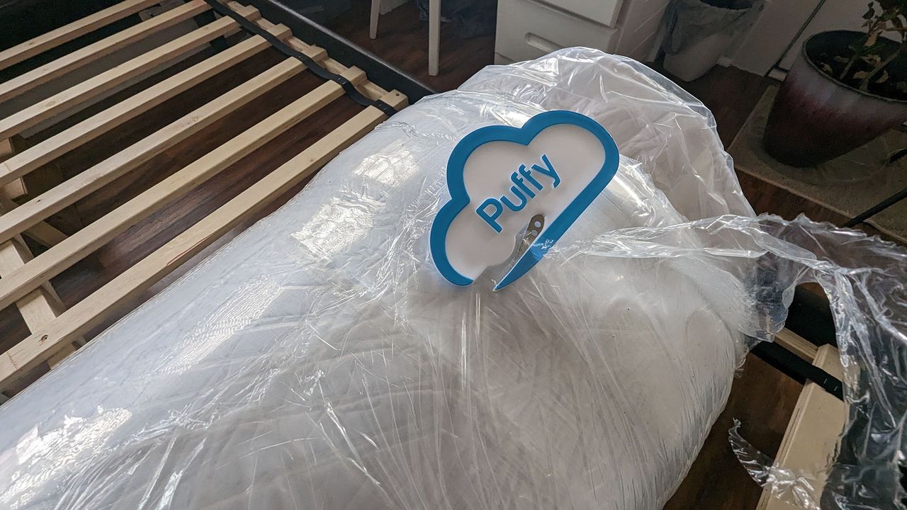 Puffy Lux Hybrid mattress vacuum packed and rolled in plastic, next to a bed frame