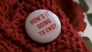 A button attached to a red sweater says 'How's It Going To End?'