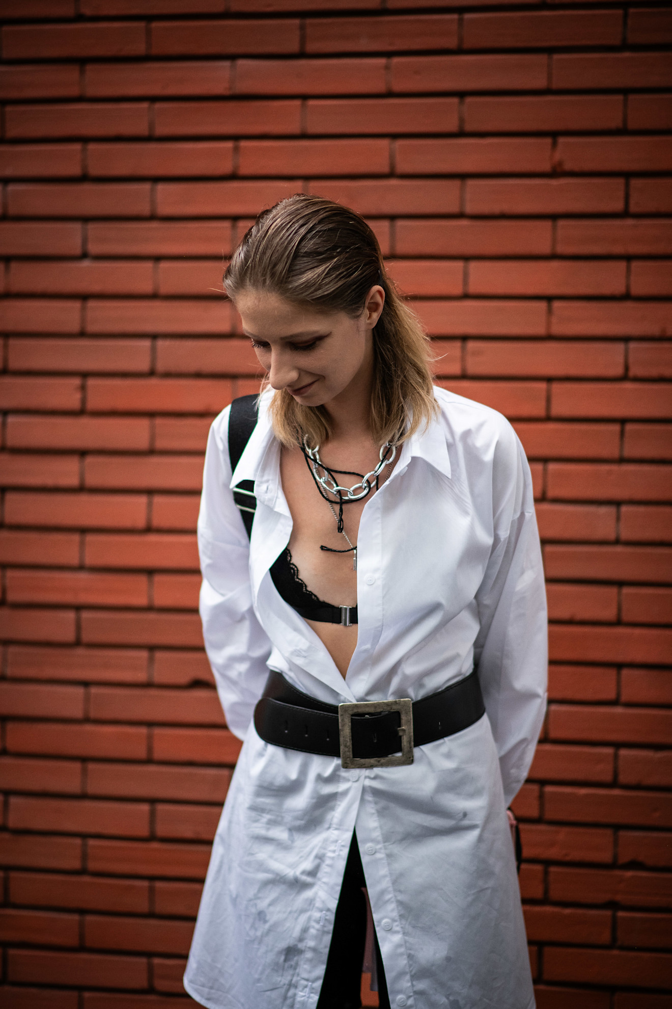 a woman with dark blonde hair is looking down wearing a white shirt, black bra and silver necklaces
