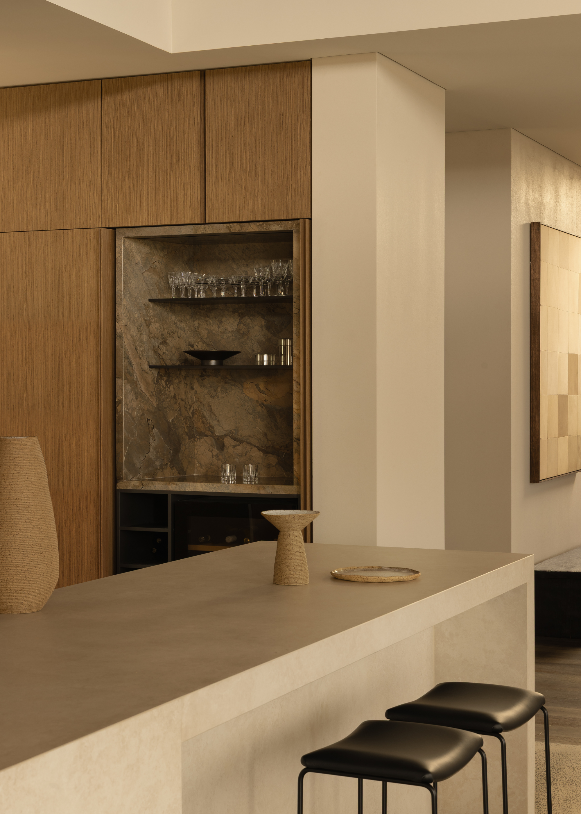 A kitchen with off-white walls, wood cabinets, an inset marble nook with floating shelves, a beige island with a pair of black stools