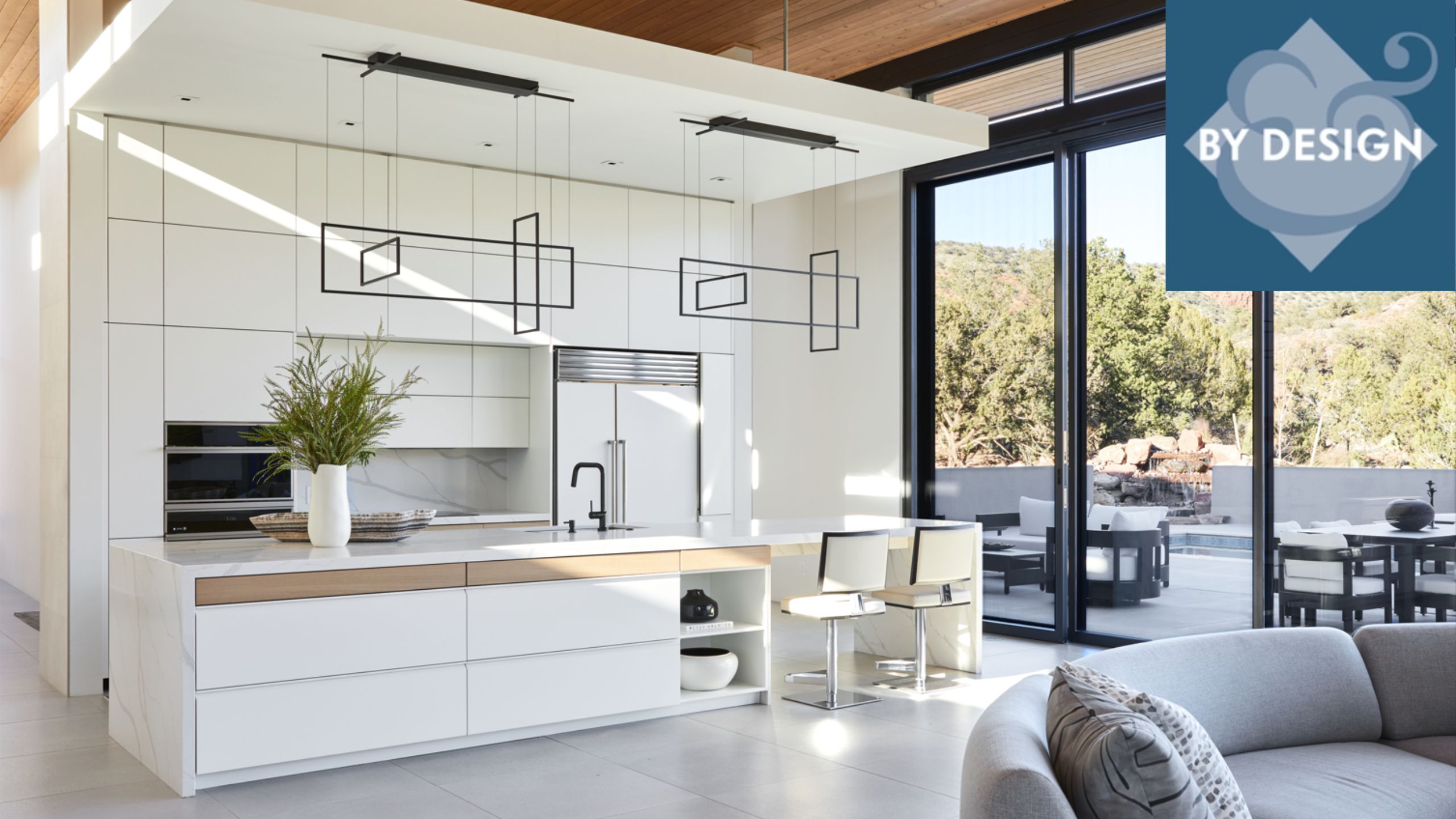 Open-plan kitchen-living space, white kitchen cabinetry, island, bar stool seating, floor-to-ceiling doors leading out to terrace, gray sofa in bottom right