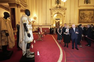 Queen Camilla and King Charles standing in a red-carpeted room in Buckingham Palace pointing at costumes from Gladiator II on display