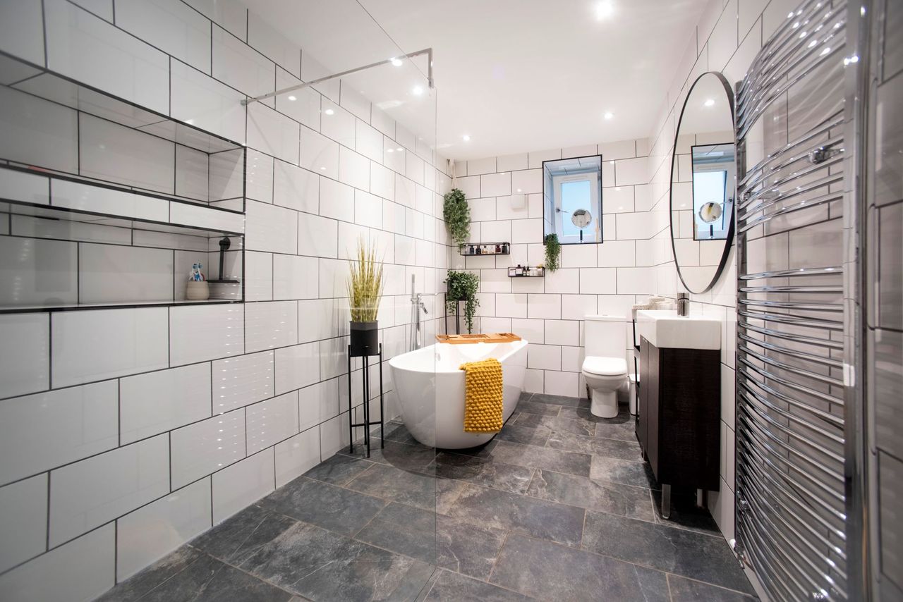 The bathroom includes white tiled walls and grey tiled floors and a white porcelain bath in the middle of the room and devils ivy on the wall