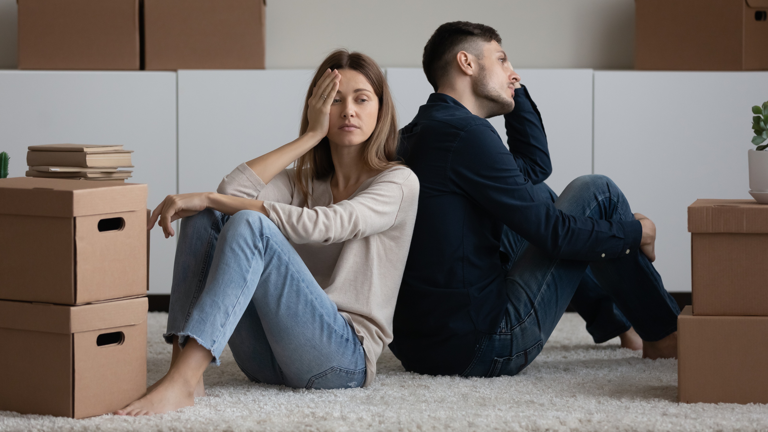 An unhappy-looking couple sit on the floor among moving boxes.