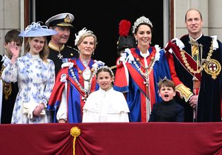 Lady Louise Windsor waving at the camera in a purple and white floral dress standing next to the Duchess of Edinburgh and Kate Middleton in blue-and-red robes, Princess Charlotte wearing a white robe and Prince Louis and Prince William dressed in black on the Buckingham Palace balcony at the coronation