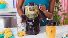 A Magic Bullet Mini Juicer making orange juice on a countertop by a woman in blue athleisure