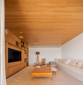 Minimalist family room with an extended beige sofa, wood furniture, wood shelving and TV on the wall.