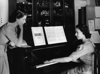 Princess Elizabeth (Queen Elizabeth II) watches her sister, Princess Margaret Countess of Snowdon (1930 - 2002) play a Brahms Waltz on the piano in the school room at Buckingham Palace.
