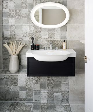 Bathroom with gray patterned tiles on the wall and floor, small white sink mounted on black wooden support, oval mirror with white frame, cream vase with dried natural grasses