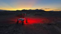 a small, four-wheeled rover shines a red light on a dark desert ground and the sun sets behind silhouetted hills in the background.