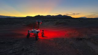 a small, four-wheeled rover shines a red light on a dark desert ground and the sun sets behind silhouetted hills in the background.