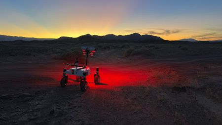 a small, four-wheeled rover shines a red light on a dark desert ground and the sun sets behind silhouetted hills in the background.