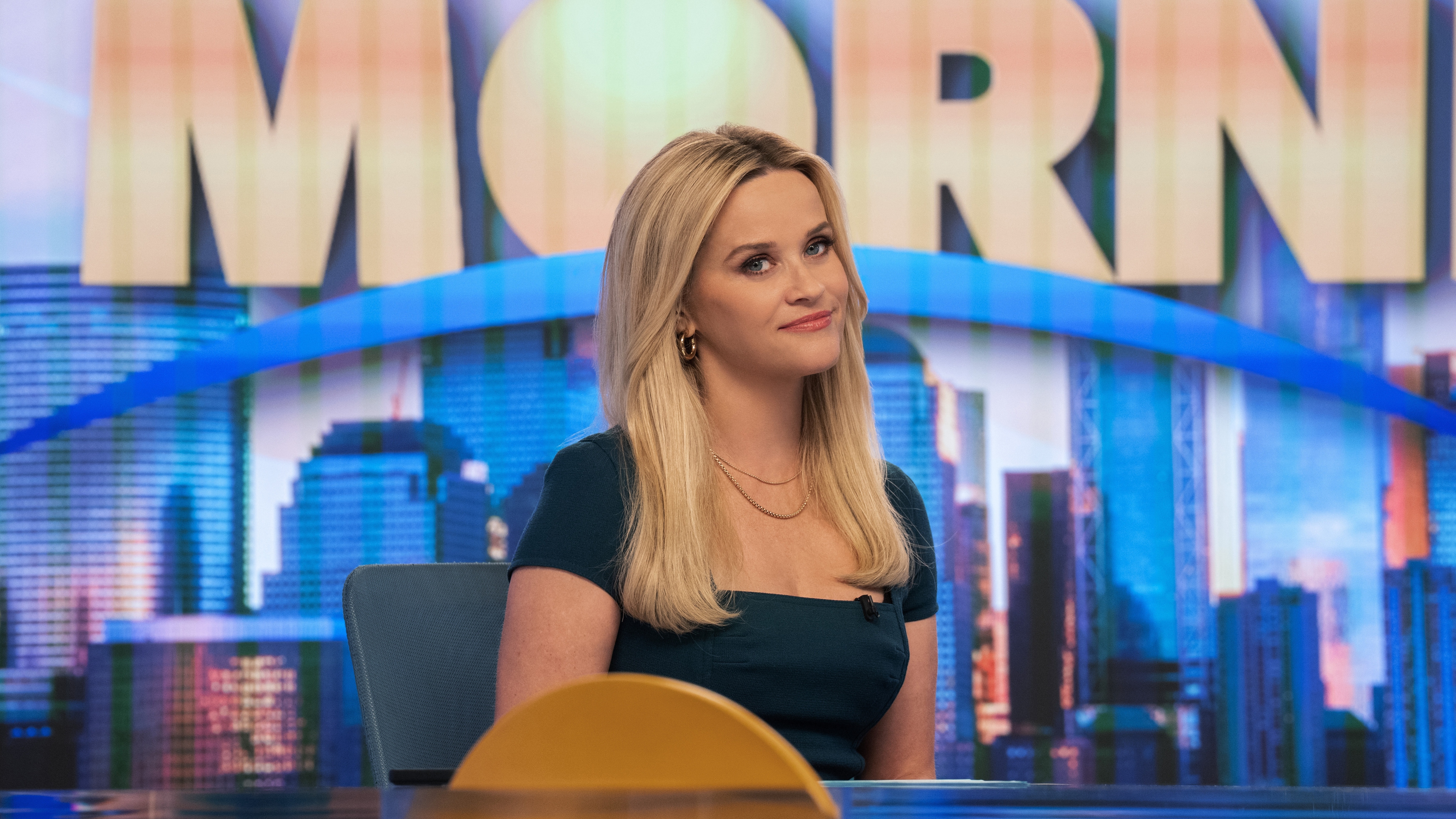 Reese Witherspoon as Bradley Jackson smiling at something off camera and sitting in front of The Morning Show sign in The Morning Show season 4.