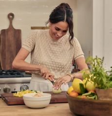 Meghan Markle wearing a beige sweater cutting lemons on a cutting board in a white kitchen