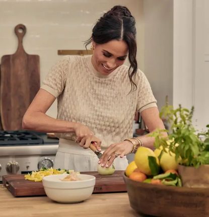 Meghan Markle wearing a beige sweater cutting lemons on a cutting board in a white kitchen