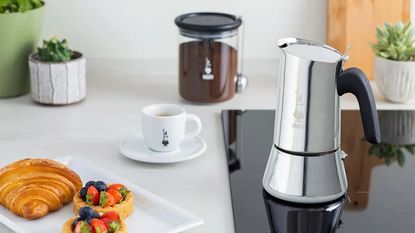 A silver Bialetti coffee pot on a black electric stove, set into a white counter. A small espresso cup beside it, with a square plate of a croissant and some flan pastries with fresh fruit.
