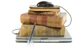 Antique books piled on a laptop computer are topped by a computer mouse.