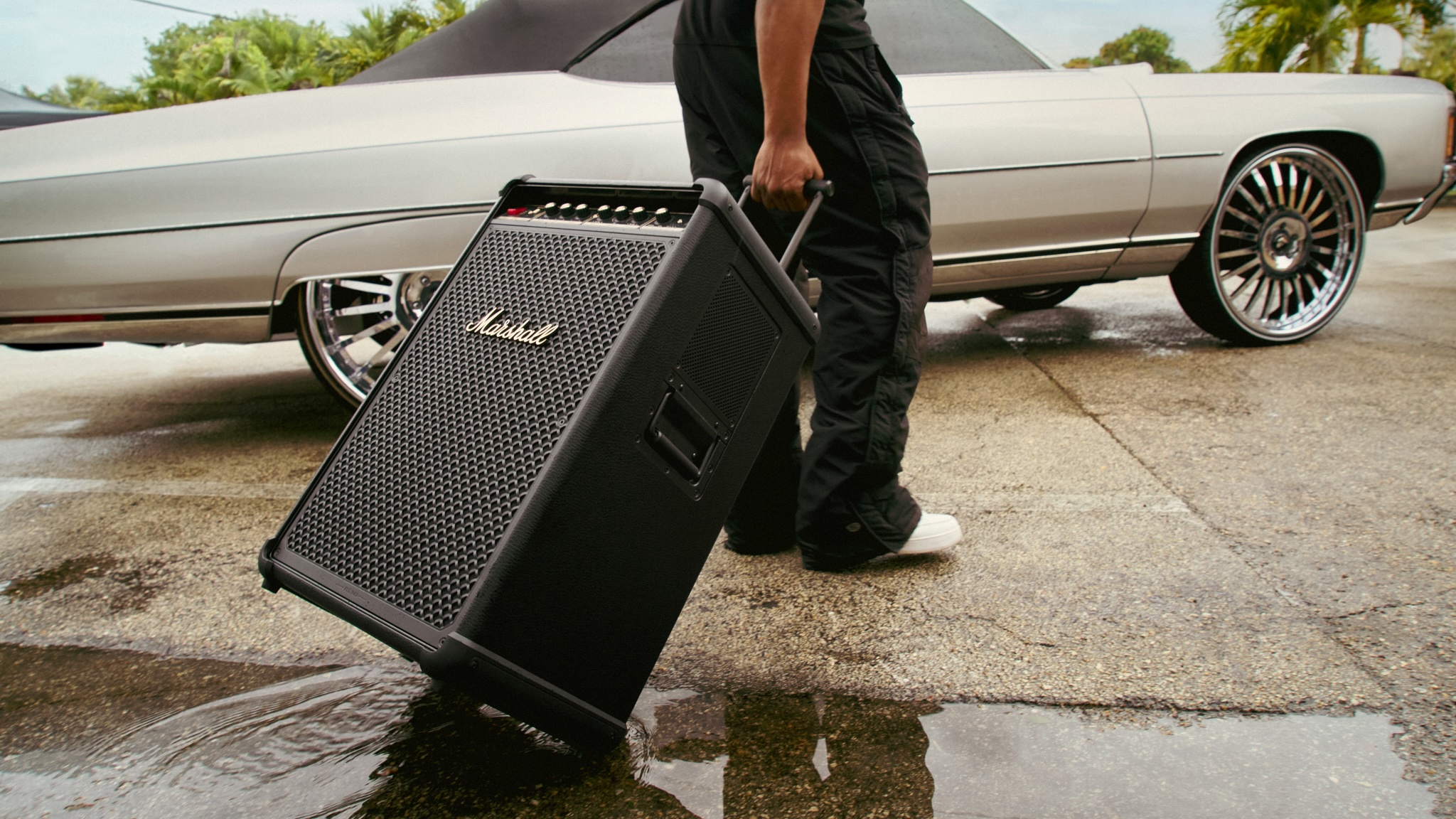 A person in dark t-shirt and combats is rolling a Marshall Bromley 750 through a puddle towards a classic American convertible car.